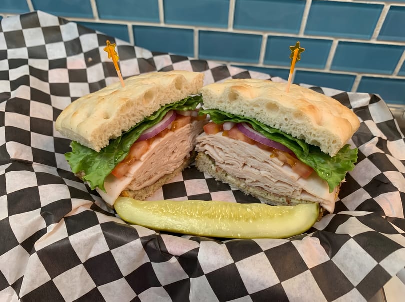 Turkey sandwich on focaccia bread with lettuce, tomato, and red onion, served with a pickle spear.