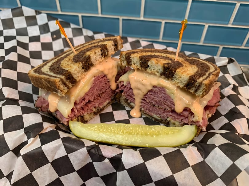 Reuben sandwich on marbled rye with melted cheese, sauce, and a pickle spear on checkered paper.