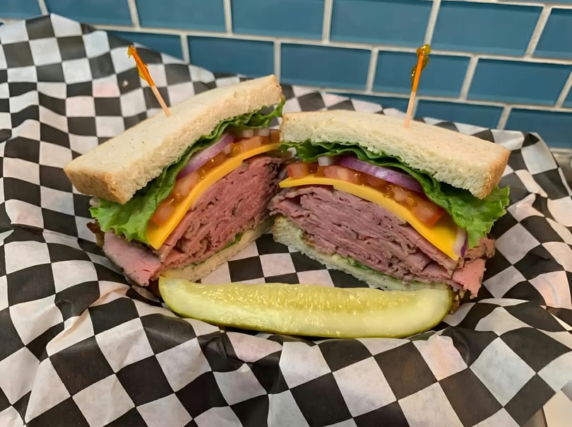 Roast beef sandwich with cheese, lettuce, tomato, and onion, served with a pickle spear.