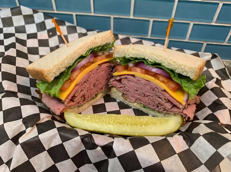 Roast beef sandwich with cheese, lettuce, tomato, and onion, served with a pickle spear.